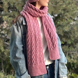 Peut inclure: Une écharpe en tricot rose avec un motif torsadé. L'écharpe est portée autour du cou d'une personne portant une veste en jean.