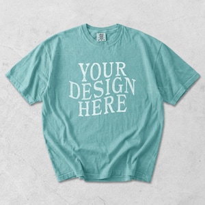 May include: A turquoise short-sleeve t-shirt with the text "YOUR DESIGN HERE" in white, centered on the front. The shirt has a crew neck and a relaxed fit. The fabric appears soft and slightly faded.