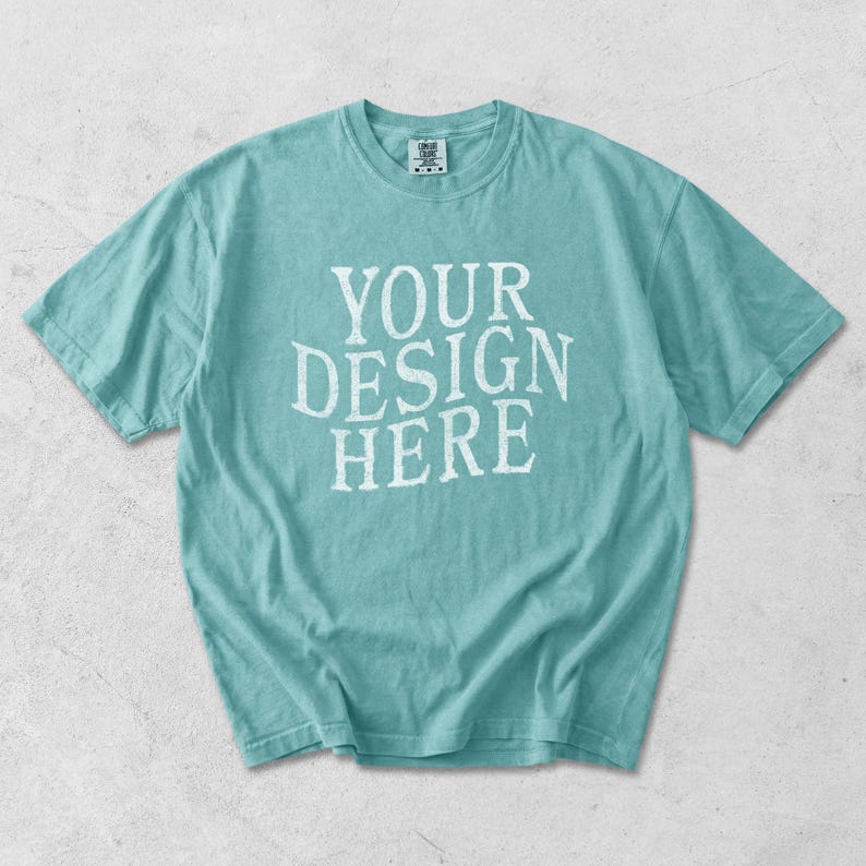 May include: A turquoise short-sleeve t-shirt with the text "YOUR DESIGN HERE" in white, centered on the front. The shirt has a crew neck and a relaxed fit. The fabric appears soft and slightly faded.