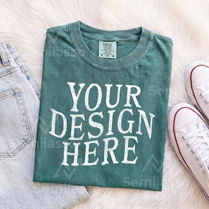 May include: A dark teal short-sleeve t-shirt with a crew neckline and a distressed white graphic print that reads YOUR DESIGN HERE. The shirt features a size M tag on the inner collar and is displayed alongside light blue denim and white sneakers.