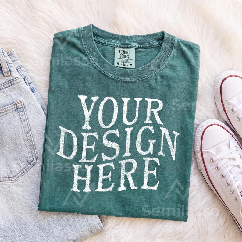 May include: A dark teal short-sleeve t-shirt with a crew neckline and a distressed white graphic print that reads YOUR DESIGN HERE. The shirt features a size M tag on the inner collar and is displayed alongside light blue denim and white sneakers.