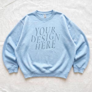 Puede incluir: Una sudadera de cuello redondo en color azul claro con el texto YOUR DESIGN HERE impreso en un tono azul m&aacute;s oscuro en el pecho. La prenda tiene mangas largas, cuello, pu&ntilde;os y dobladillo acanalados, con una etiqueta de la marca Gildan en el cuello.