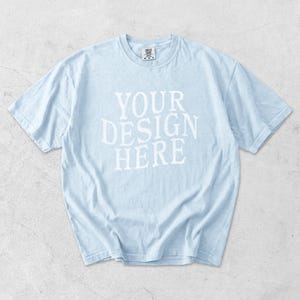 May include: Light blue t-shirt with the text "YOUR DESIGN HERE" in white block letters. The shirt has short sleeves and a crew neck. The fabric appears soft and slightly wrinkled.