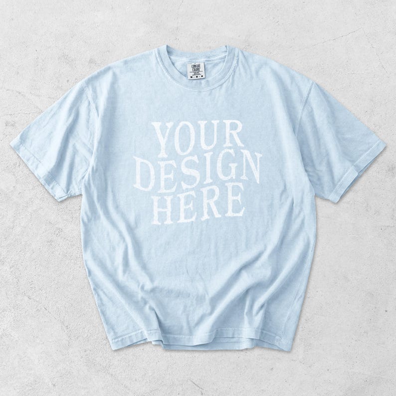 May include: Light blue t-shirt with the text "YOUR DESIGN HERE" in white block letters. The shirt has short sleeves and a crew neck. The fabric appears soft and slightly wrinkled.
