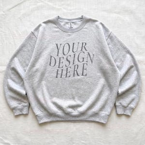 Puede incluir: Una sudadera de cuello redondo en color gris jaspeado claro con el texto gris oscuro "YOUR DESIGN HERE" impreso en el pecho en una fuente serif centrada y ligeramente arqueada. La prenda tiene mangas largas, pu&ntilde;os y cuello acanalados.