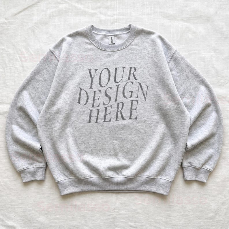 Puede incluir: Una sudadera de cuello redondo en color gris jaspeado claro con el texto gris oscuro "YOUR DESIGN HERE" impreso en el pecho en una fuente serif centrada y ligeramente arqueada. La prenda tiene mangas largas, pu&ntilde;os y cuello acanalados.