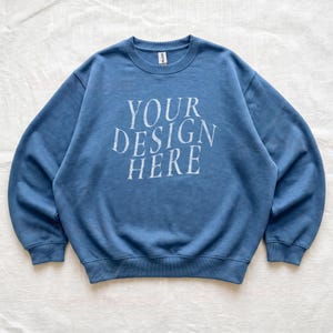 Puede incluir: Una sudadera de cuello redondo en color azul polvoriento con el texto blanco "YOUR DESIGN HERE" impreso en una fuente serif desgastada en el pecho. La prenda tiene mangas largas con pu&ntilde;os acanalados, cuello acanalado y dobladillo acanalado.