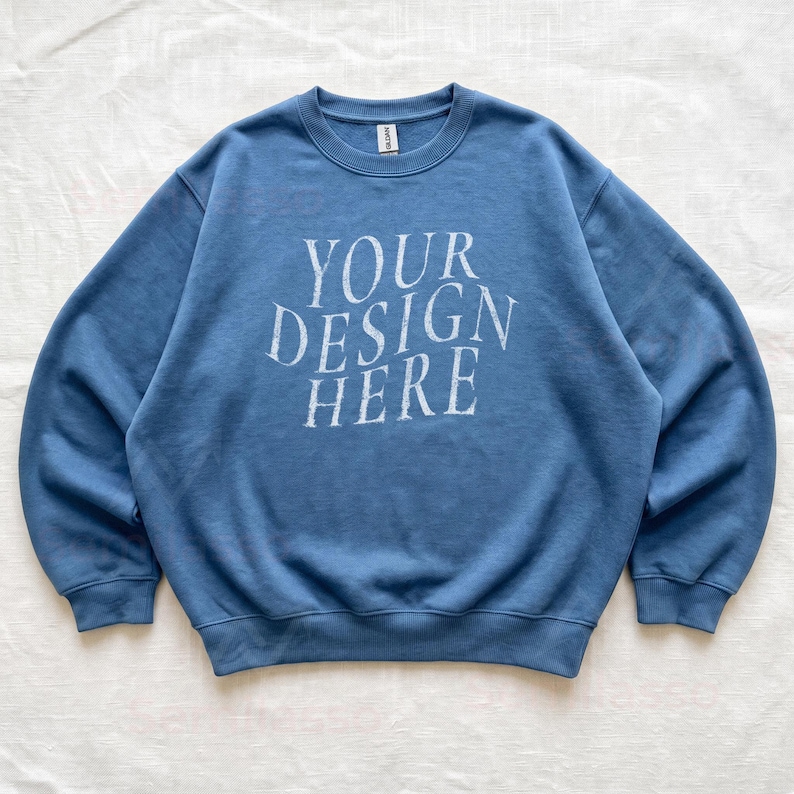 Puede incluir: Una sudadera de cuello redondo en color azul polvoriento con el texto blanco "YOUR DESIGN HERE" impreso en una fuente serif desgastada en el pecho. La prenda tiene mangas largas con pu&ntilde;os acanalados, cuello acanalado y dobladillo acanalado.