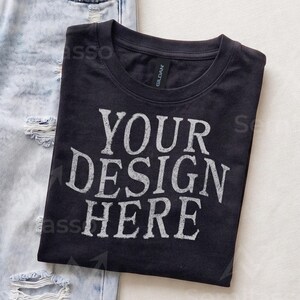 May include: A black crew neck t-shirt featuring the white distressed text "YOUR DESIGN HERE" printed on the front. The shirt has a Gildan brand label at the inner neckline and is positioned next to a pair of light blue distressed denim jeans.