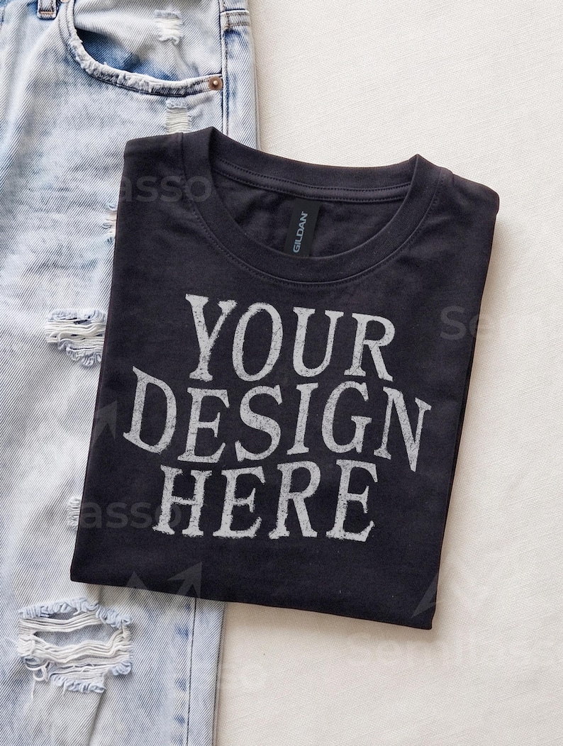 May include: A black crew neck t-shirt featuring the white distressed text "YOUR DESIGN HERE" printed on the front. The shirt has a Gildan brand label at the inner neckline and is positioned next to a pair of light blue distressed denim jeans.