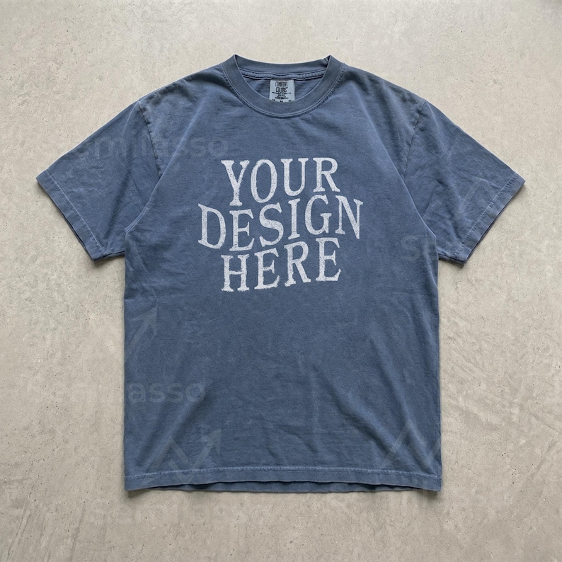 May include: A slate blue short-sleeve t-shirt with a crew neckline. The center of the chest features the text "YOUR DESIGN HERE" printed in a distressed, off-white, serif font. The fabric has a washed, vintage appearance with visible seams.
