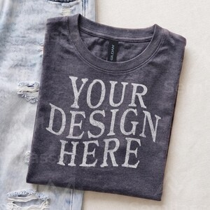 May include: A charcoal gray crew neck t-shirt with the white text "YOUR DESIGN HERE" printed on the front. The shirt features a Gildan brand label at the inner neckline and is positioned next to a pair of light blue distressed denim jeans.