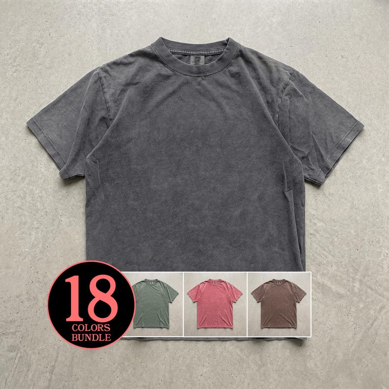 May include: A charcoal grey short-sleeve t-shirt with a crew neckline and a washed-out texture. A circular graphic in the bottom left corner displays the text 18 COLORS BUNDLE, featuring small images of green, pink, and brown t-shirts below it.