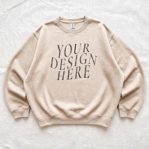 Puede incluir: Una sudadera beige de cuello redondo con el texto YOUR DESIGN HERE impreso en una fuente gris oscuro desgastada en el pecho. La prenda tiene mangas largas, cuello, pu&ntilde;os y dobladillo acanalados, junto con una etiqueta de marca visible.