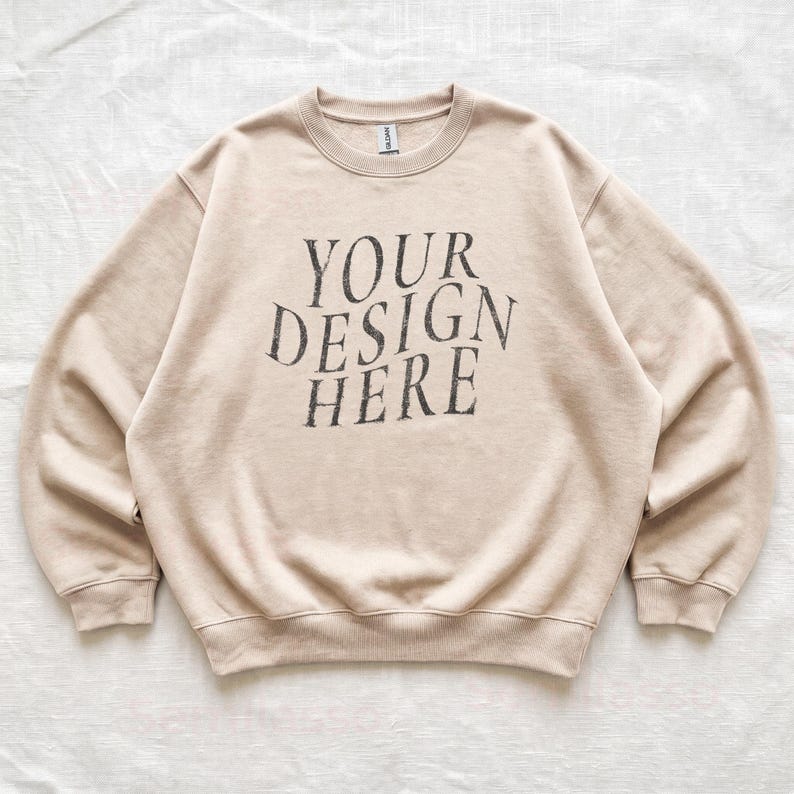 Puede incluir: Una sudadera beige de cuello redondo con el texto YOUR DESIGN HERE impreso en una fuente gris oscuro desgastada en el pecho. La prenda tiene mangas largas, cuello, pu&ntilde;os y dobladillo acanalados, junto con una etiqueta de marca visible.