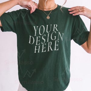 Può includere: Una t-shirt a maniche corte verde bosco scuro con la scritta bianca "YOUR DESIGN HERE" stampata in un carattere serif usurato sul petto. Il capo presenta un girocollo e una vestibilità rilassata, indossato con pantaloncini di colore chiaro.