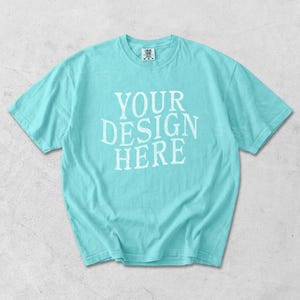 May include: A light turquoise t-shirt with the text "YOUR DESIGN HERE" in white, centered on the front. The short-sleeved shirt is made of a soft material and is laid flat on a light gray surface.