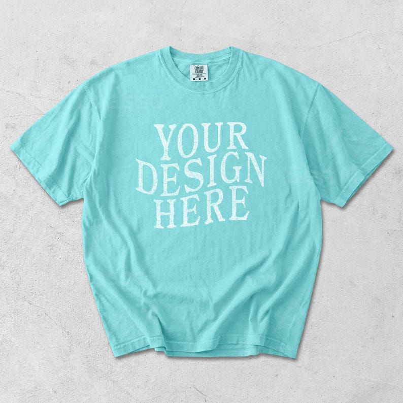 May include: A light turquoise t-shirt with the text "YOUR DESIGN HERE" in white, centered on the front. The short-sleeved shirt is made of a soft material and is laid flat on a light gray surface.