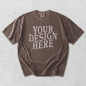 May include: A brown t-shirt with the text "YOUR DESIGN HERE" in white, vintage-style lettering. The shirt has a crew neck and short sleeves. The fabric appears to be pre-shrunk and has a slightly faded look.