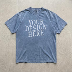 May include: A short-sleeve crew neck t-shirt in a faded blue wash with a distressed texture. The center of the chest features the white text "YOUR DESIGN HERE" printed in a bold, serif font. The garment has a standard fit with visible stitching.