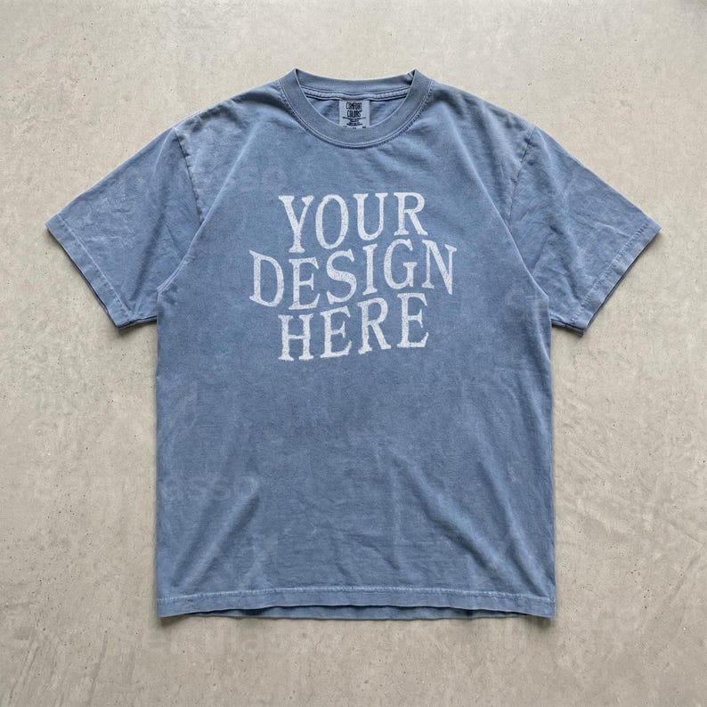 May include: A short-sleeve crew neck t-shirt in a faded blue wash with a distressed texture. The center of the chest features the white text "YOUR DESIGN HERE" printed in a bold, serif font. The garment has a standard fit with visible stitching.