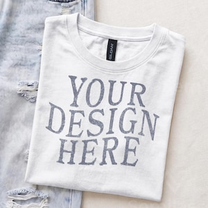 May include: A white crew neck t-shirt with the text YOUR DESIGN HERE printed in a distressed grey font on the chest. The shirt features a black Gildan brand label at the inner neckline and is positioned next to a pair of light blue distressed denim jeans.