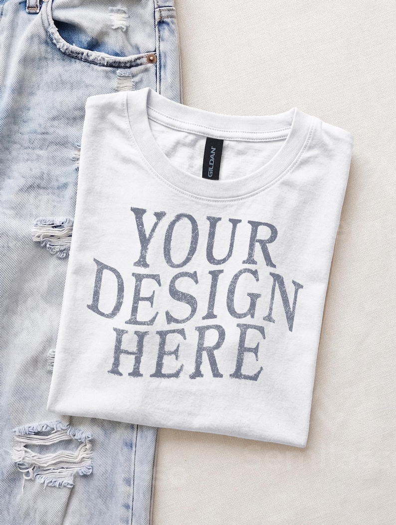 May include: A white crew neck t-shirt with the text YOUR DESIGN HERE printed in a distressed grey font on the chest. The shirt features a black Gildan brand label at the inner neckline and is positioned next to a pair of light blue distressed denim jeans.