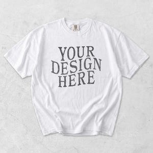 May include: White t-shirt with the text "YOUR DESIGN HERE" in a distressed, dark gray font. The shirt has a crew neck and short sleeves. The fabric appears to be soft and slightly wrinkled.