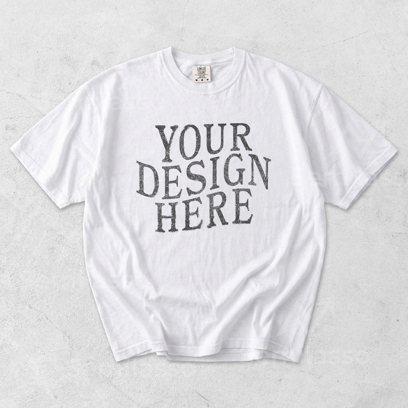 May include: White t-shirt with the text "YOUR DESIGN HERE" in a distressed, dark gray font. The shirt has a crew neck and short sleeves. The fabric appears to be soft and slightly wrinkled.