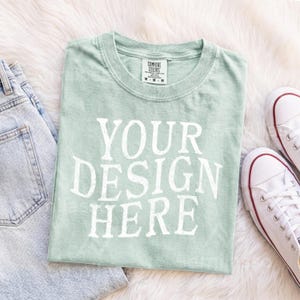 May include: A light sage green short-sleeve t-shirt with a crew neckline and the white text "YOUR DESIGN HERE" printed on the front. The shirt features a Comfort Colors brand label on the inside collar indicating a size medium.