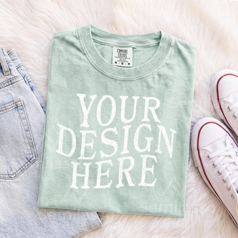 May include: A light sage green short-sleeve t-shirt with a crew neckline and the white text "YOUR DESIGN HERE" printed on the front. The shirt features a Comfort Colors brand label on the inside collar indicating a size medium.