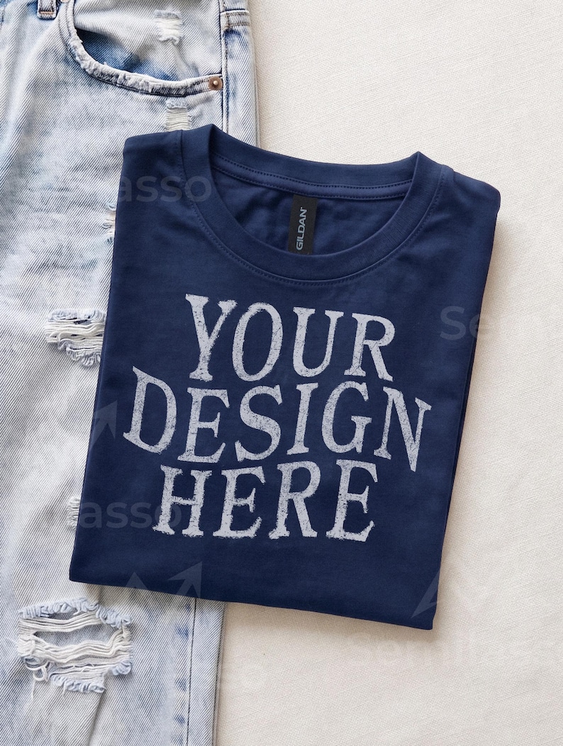 May include: A navy blue crew neck t-shirt featuring the white distressed text "YOUR DESIGN HERE" printed on the front. The shirt has a Gildan brand label at the inner neckline and is positioned next to a pair of light blue distressed denim jeans.