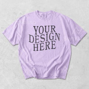 May include: A light purple t-shirt with the text "YOUR DESIGN HERE" in a distressed black font. The shirt is a crew neck style and appears to be made of a soft material. The shirt is laid flat on a light gray surface.