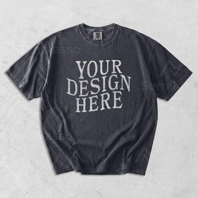 May include: A dark gray t-shirt with the text "YOUR DESIGN HERE" in a distressed white font. The shirt has a crew neck and short sleeves. The fabric appears to be pre-washed for a vintage look.