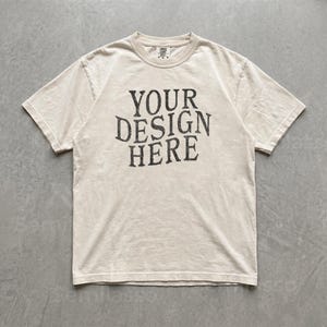 May include: A short-sleeve crew neck t-shirt in a light beige color. The center of the chest features the text "YOUR DESIGN HERE" printed in a dark charcoal gray, distressed, serif font. The garment has a standard fit with visible stitching.