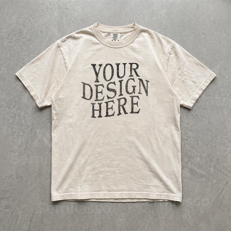 May include: A short-sleeve crew neck t-shirt in a light beige color. The center of the chest features the text "YOUR DESIGN HERE" printed in a dark charcoal gray, distressed, serif font. The garment has a standard fit with visible stitching.