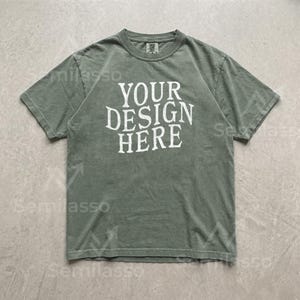 May include: A sage green short-sleeve t-shirt with a crew neckline. The center of the chest features the white text "YOUR DESIGN HERE" in a distressed, serif font. The garment has a standard fit with visible stitching along the sleeves and hem.