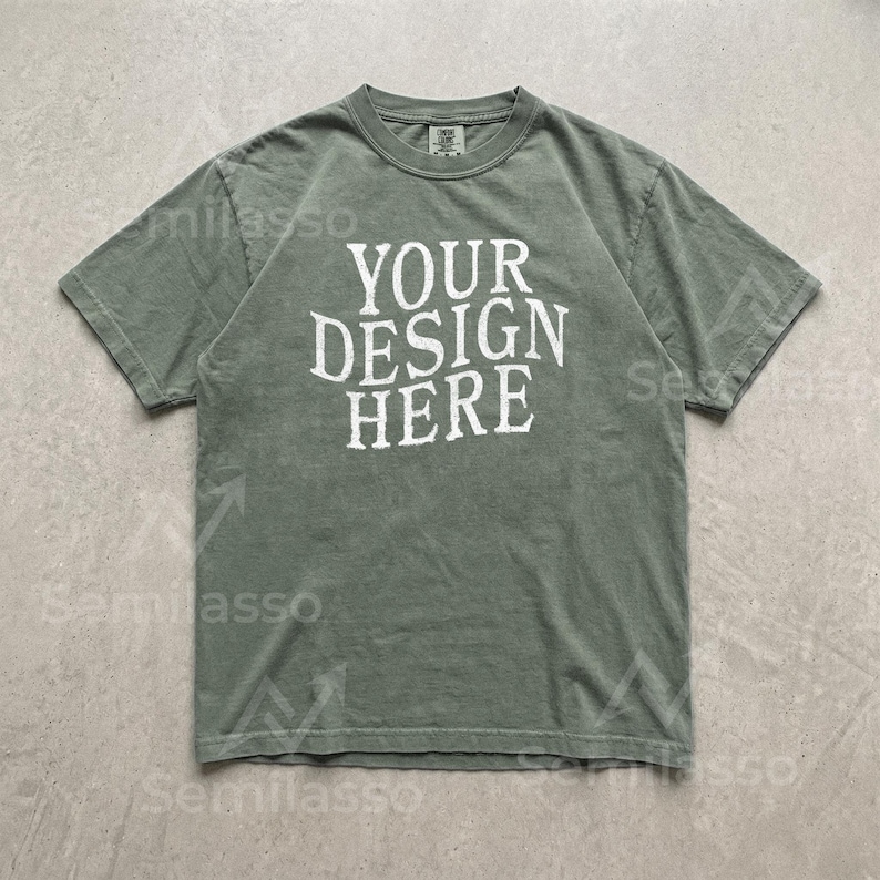 May include: A sage green short-sleeve t-shirt with a crew neckline. The center of the chest features the white text "YOUR DESIGN HERE" in a distressed, serif font. The garment has a standard fit with visible stitching along the sleeves and hem.