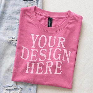 May include: A pink short-sleeved t-shirt with a crew neckline and the white text "YOUR DESIGN HERE" printed on the front. The shirt features a black Gildan brand label at the inner neck and is positioned next to light blue distressed denim jeans.