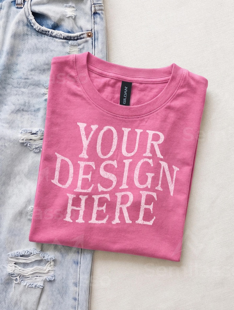 May include: A pink short-sleeved t-shirt with a crew neckline and the white text "YOUR DESIGN HERE" printed on the front. The shirt features a black Gildan brand label at the inner neck and is positioned next to light blue distressed denim jeans.
