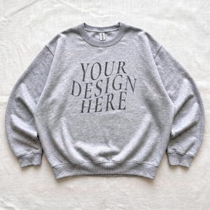 Puede incluir: Una sudadera de cuello redondo en color gris claro con el texto "YOUR DESIGN HERE" en gris oscuro impreso en una fuente serif en el pecho. La prenda tiene mangas largas, cuello, pu&ntilde;os y dobladillo acanalados, con una etiqueta de marca visible.