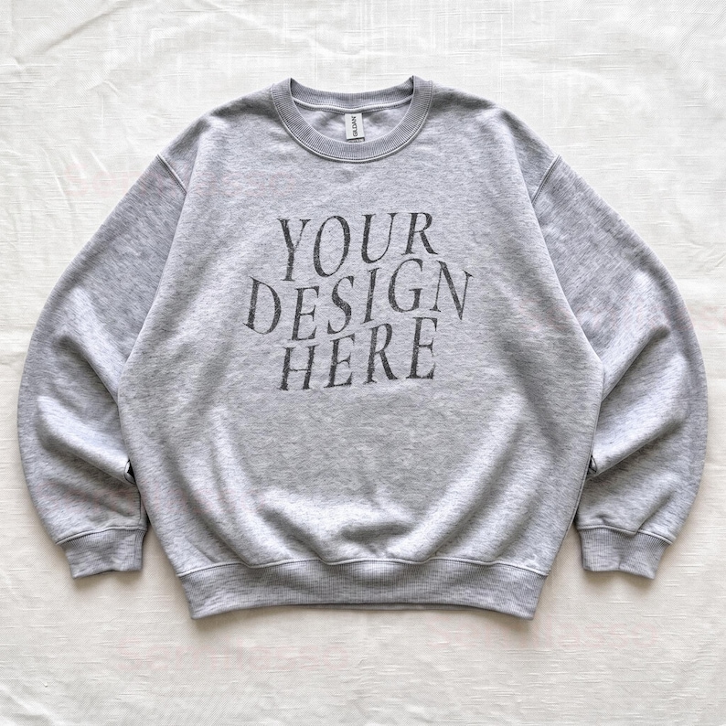 Puede incluir: Una sudadera de cuello redondo en color gris claro con el texto "YOUR DESIGN HERE" en gris oscuro impreso en una fuente serif en el pecho. La prenda tiene mangas largas, cuello, pu&ntilde;os y dobladillo acanalados, con una etiqueta de marca visible.