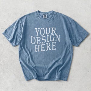 May include: A faded blue t-shirt with the text "YOUR DESIGN HERE" in white block letters. The shirt has a crew neck and short sleeves. The fabric appears soft and slightly worn.