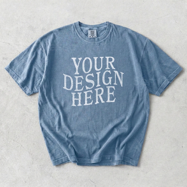 May include: A faded blue t-shirt with the text "YOUR DESIGN HERE" in white block letters. The shirt has a crew neck and short sleeves. The fabric appears soft and slightly worn.