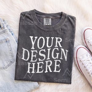 May include: A charcoal gray short-sleeve t-shirt with a crew neckline and the white distressed-style text "YOUR DESIGN HERE" printed on the front. The shirt features a size M tag from Comfort Colors and is made of 100% cotton material.