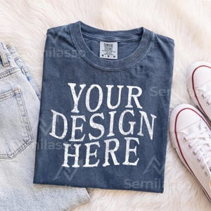 May include: A dark blue short-sleeved t-shirt with a crew neckline featuring the white text "YOUR DESIGN HERE" printed in a bold, distressed font on the front. The shirt includes a white interior neck label with black text and size M.