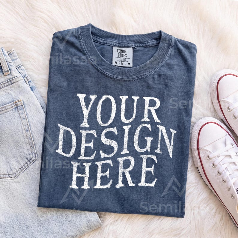 May include: A dark blue short-sleeved t-shirt with a crew neckline featuring the white text "YOUR DESIGN HERE" printed in a bold, distressed font on the front. The shirt includes a white interior neck label with black text and size M.