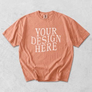 May include: A peach-colored t-shirt with the text "YOUR DESIGN HERE" in white, distressed lettering. The short-sleeved shirt has a crew neck and a slightly faded appearance, suggesting a vintage or worn look. The shirt is a blank canvas for custom designs.