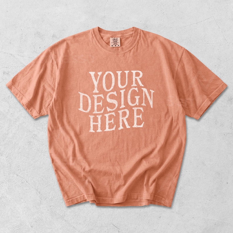 May include: A peach-colored t-shirt with the text "YOUR DESIGN HERE" in white, distressed lettering. The short-sleeved shirt has a crew neck and a slightly faded appearance, suggesting a vintage or worn look. The shirt is a blank canvas for custom designs.