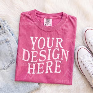 May include: A pink short-sleeved t-shirt with a crew neckline featuring the white text YOUR DESIGN HERE printed in a bold, sans-serif font across the chest. The shirt has a visible interior neck label with the text COMFORT COLORS size M-M.
